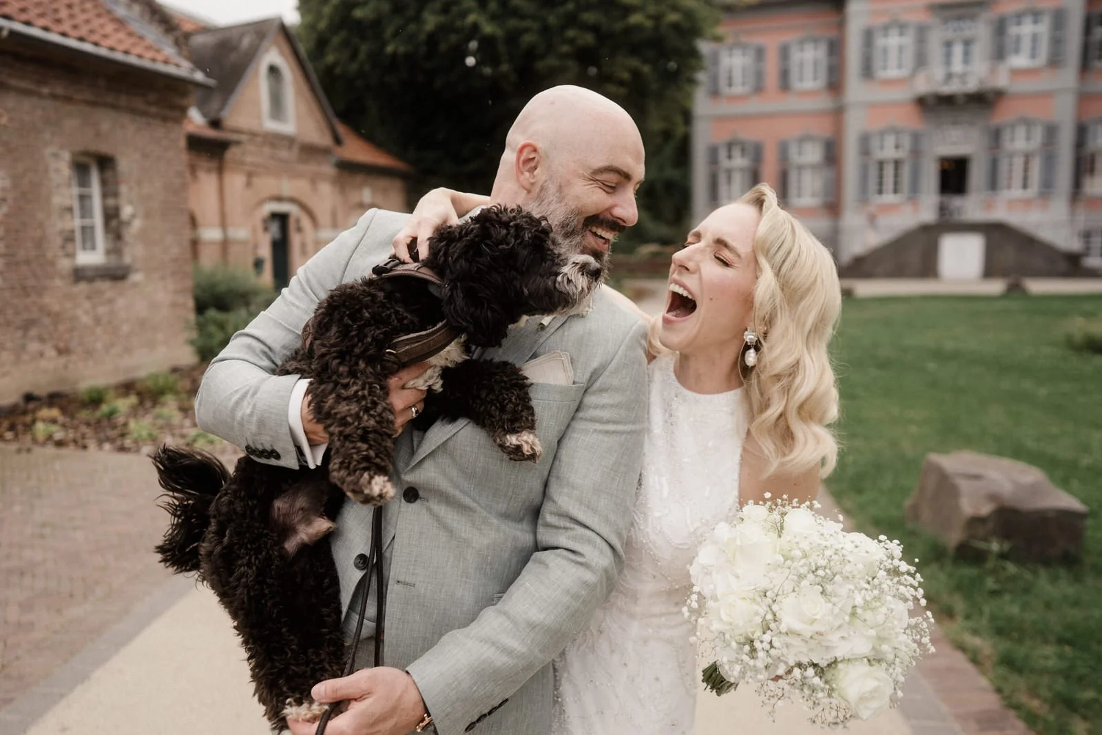 Ein Mann in einem grauen Anzug, eine Frau in einem weißen Brautkleid, lachen zusammen, während der Mann einen schwarzen Hund hält, im Freien mit historischen Gebäuden im Hintergrund.