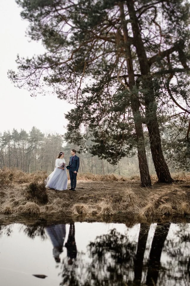 Ein Brautpaar steht am Ufer eines Sees in einem Wald, mit Blick auf das Wasser, umgeben von Bäumen und Natur.