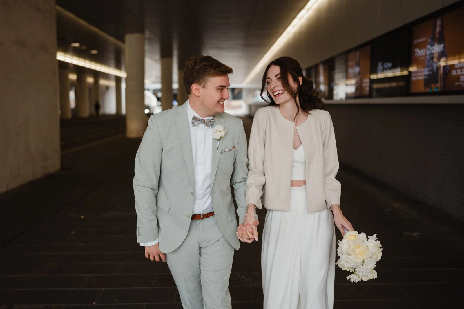 Ein lachendes Paar, vermutlich frisch verheiratet, das Hand in Hand durch einen Tunnel oder eine Unterführung geht, die Frau hält einen Brautstrauß in der Hand.
