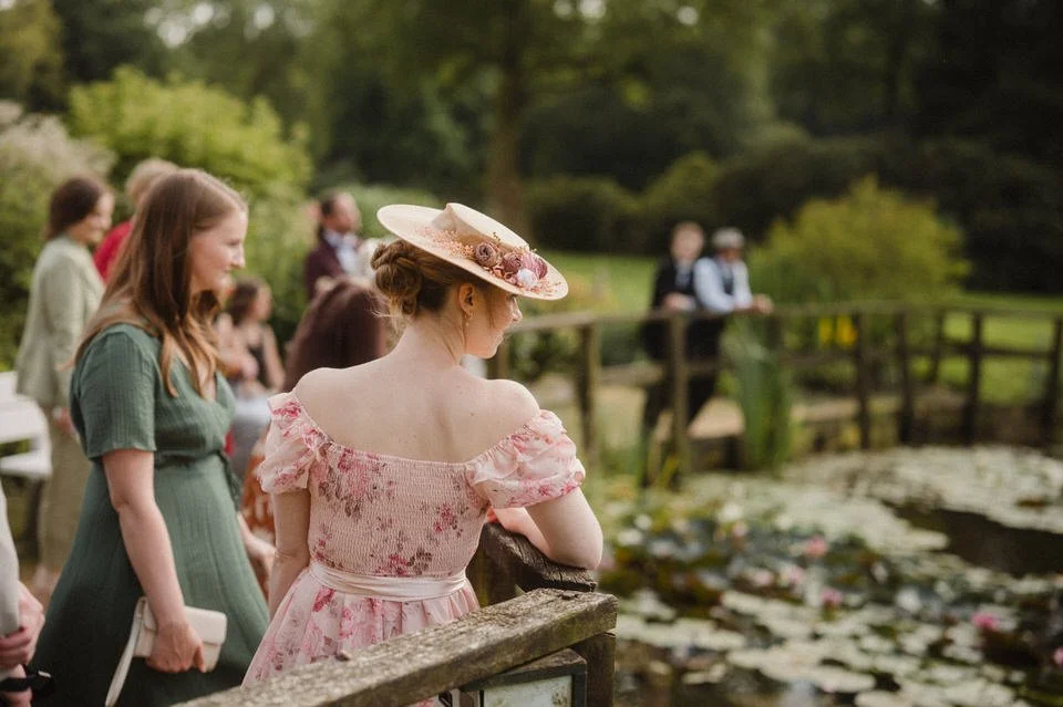 Menschen, die an einem Teich mit Seerosen in einem Park spazieren gehen, viele tragen historische Kleidung, eine Frau trägt einen Strohhut.