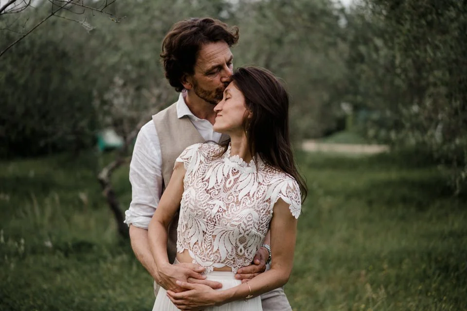 Ein Paar steht im Grünen, der Mann küsst die Frau auf die Stirn, sie umarmen sich liebevoll.