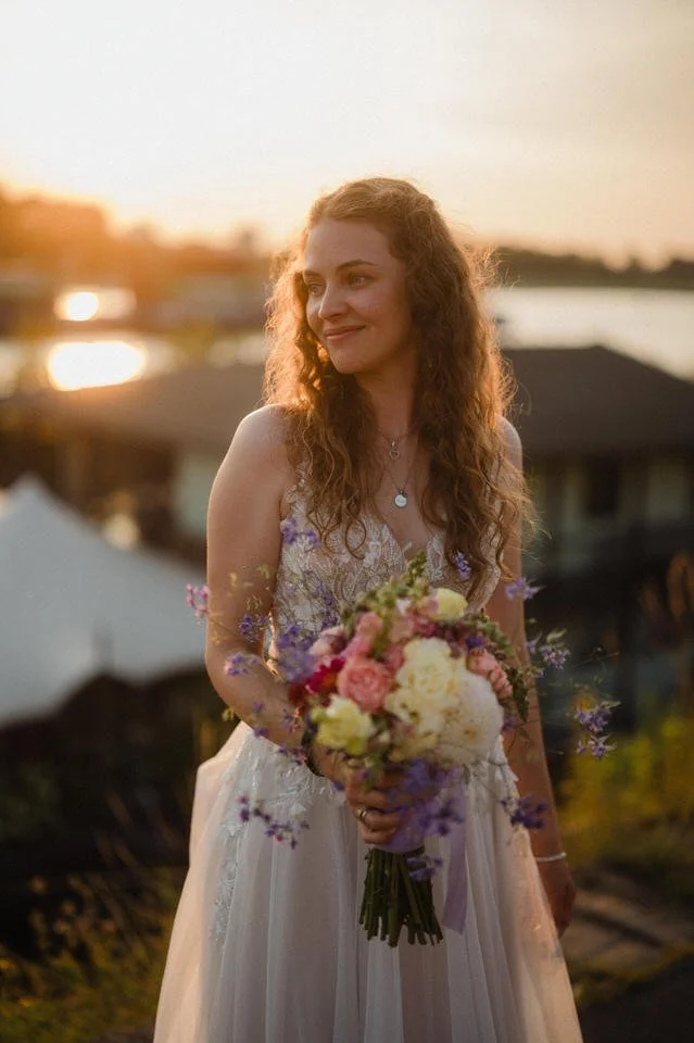 Junge Frau trägt ein weißes Brautkleid und hält einen Blumenstrauß während des Sonnenuntergangs im Freien.