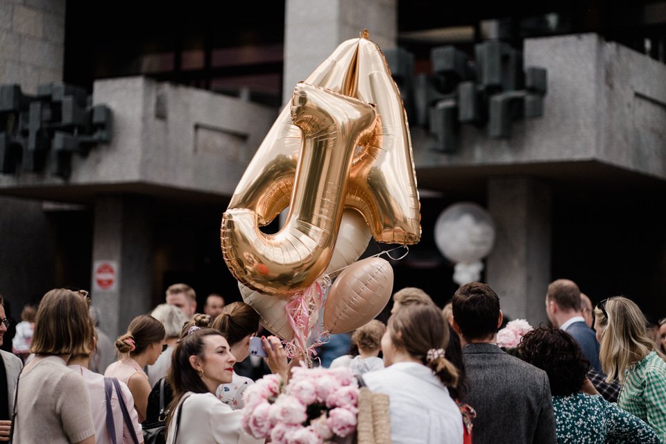 Menschen versammeln sich bei einer Feier, im Mittelpunkt ein großer, goldener Luftballon in Form einer Zahl sieben.