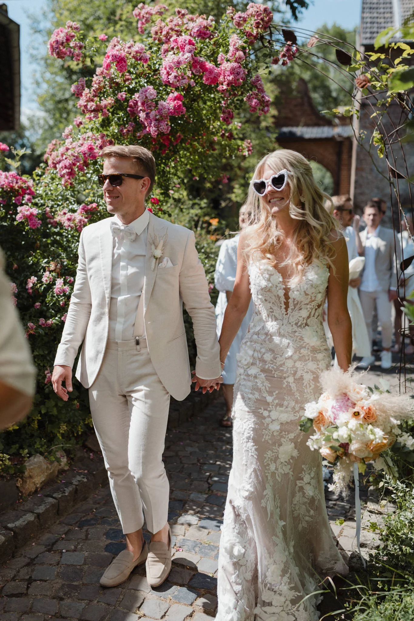 Braut und Bräutigam beim Heiratsfest, die Braut trägt eine weiße Hochzeitskleid mit Spitze und Sonnenbrille, der Bräutigam trägt einen hellen Anzug und Sonnenbrille, im Hintergrund sind Hochzeitsgäste und bunte Blumen. Es ist ein sonniger Tag im Freien.