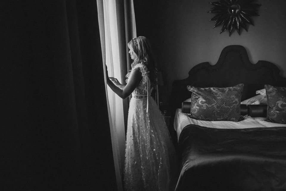 Junge Frau in einem weißen Kleid steht neben einem Fenster und liest eine Nachricht auf ihrem Handy in einem Schlafzimmer mit bett, Kissen und dunklem Kopfteil, in Schwarz-Weiß-Fotografie.