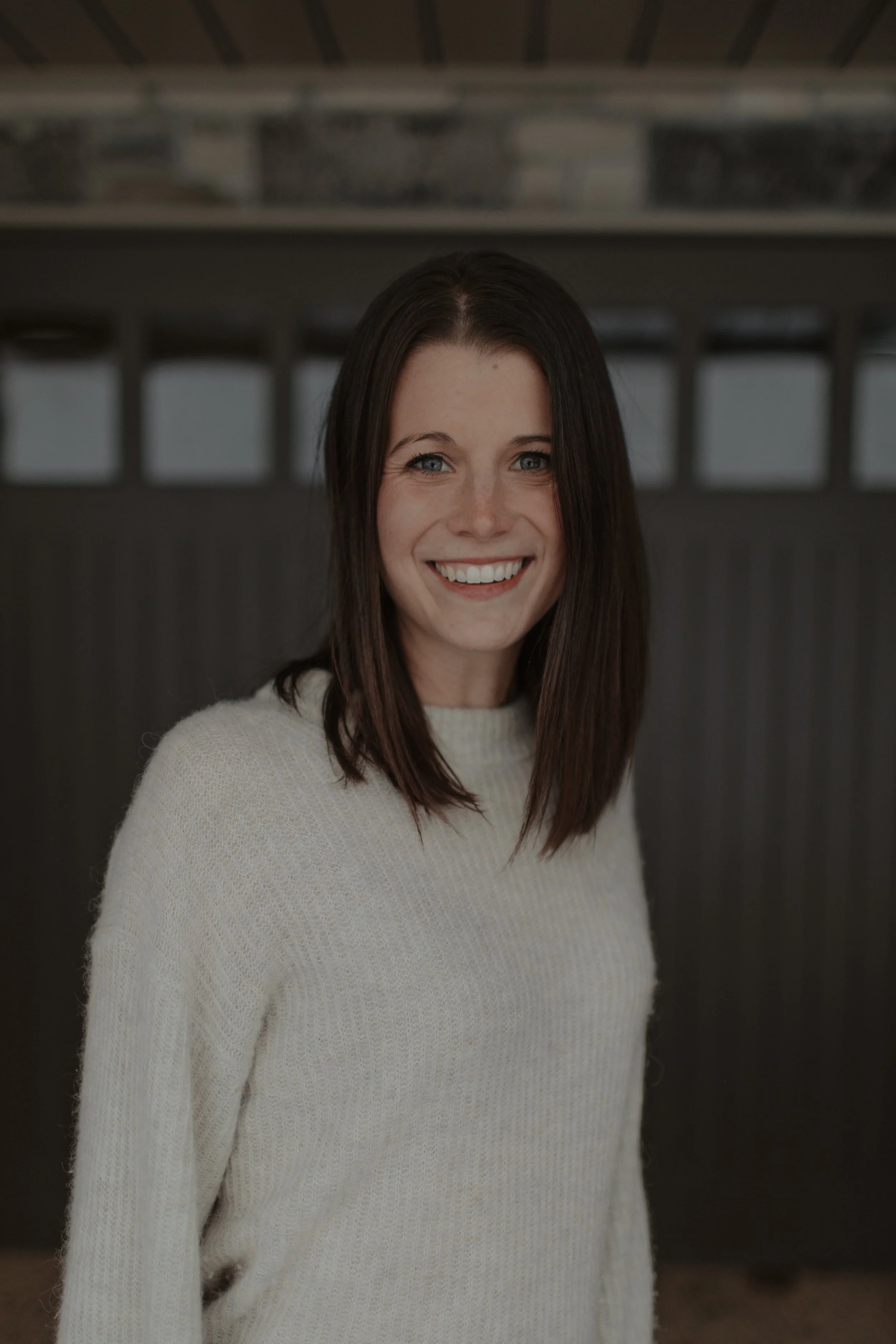 A young woman with straight, shoulder-length brown hair and blue eyes, smiling at the camera, wearing a cream-colored sweater, standing indoors with a blurred background.