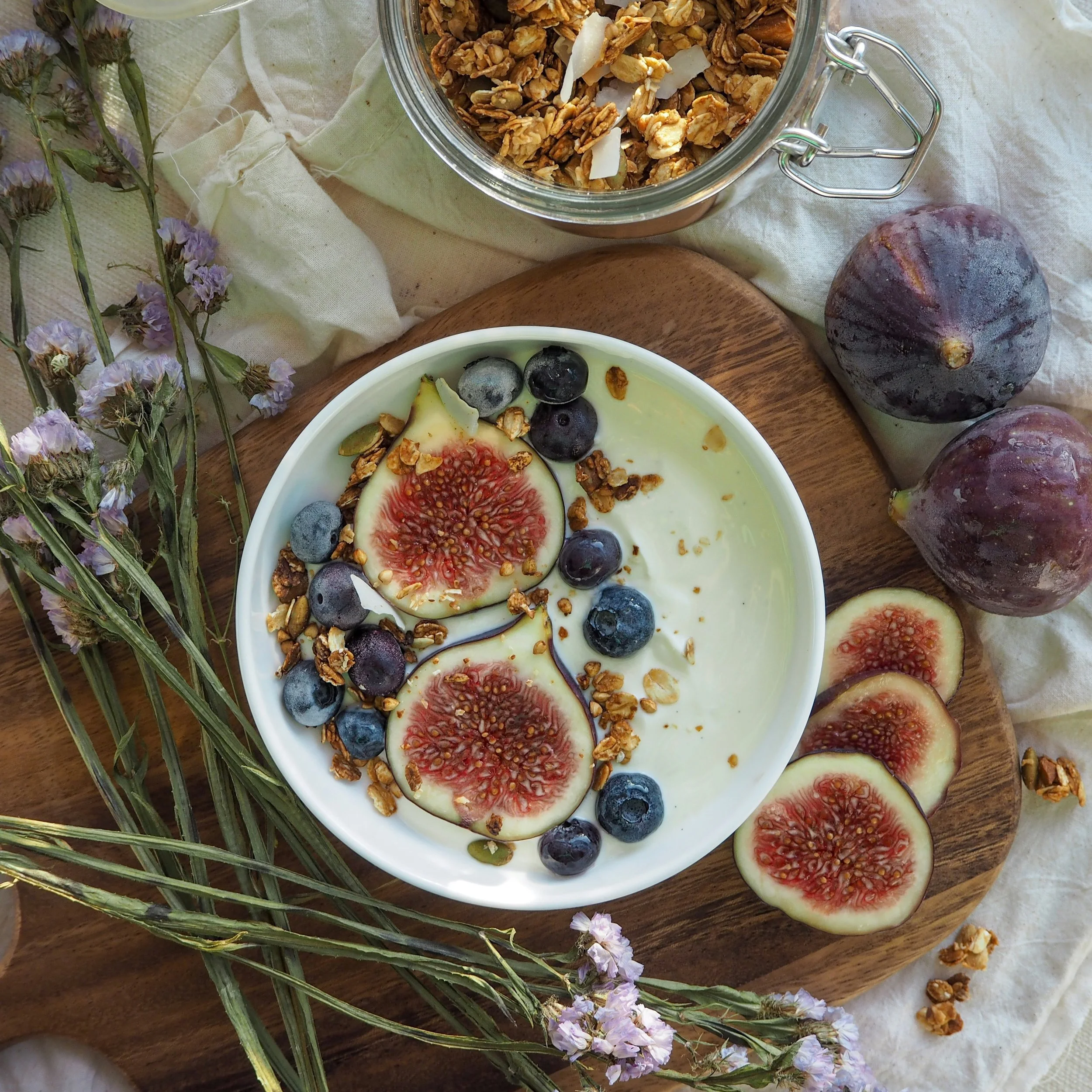Yogurt topped with sliced figs, blueberries, granola, and coconut flakes served in a white bowl on a wooden platter, with additional figs and granola nearby, and dried flowers alongside.