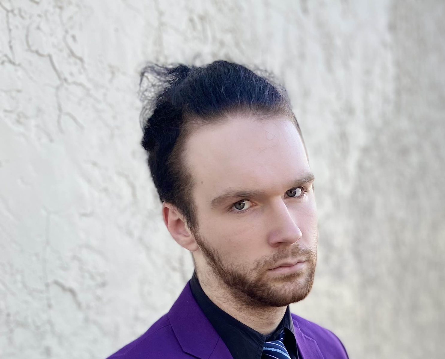 A young man with stylish dark hair, a beard, and blue eyes, wearing a purple jacket and a striped tie, posing against a textured off-white wall.