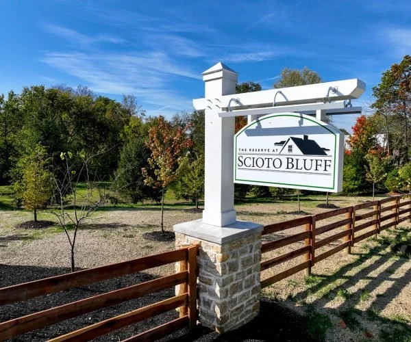 Sign for Scioto Bluff estate, mounted on a white post with a brick base, in front of a wooden fence, with trees and a blue sky in the background.