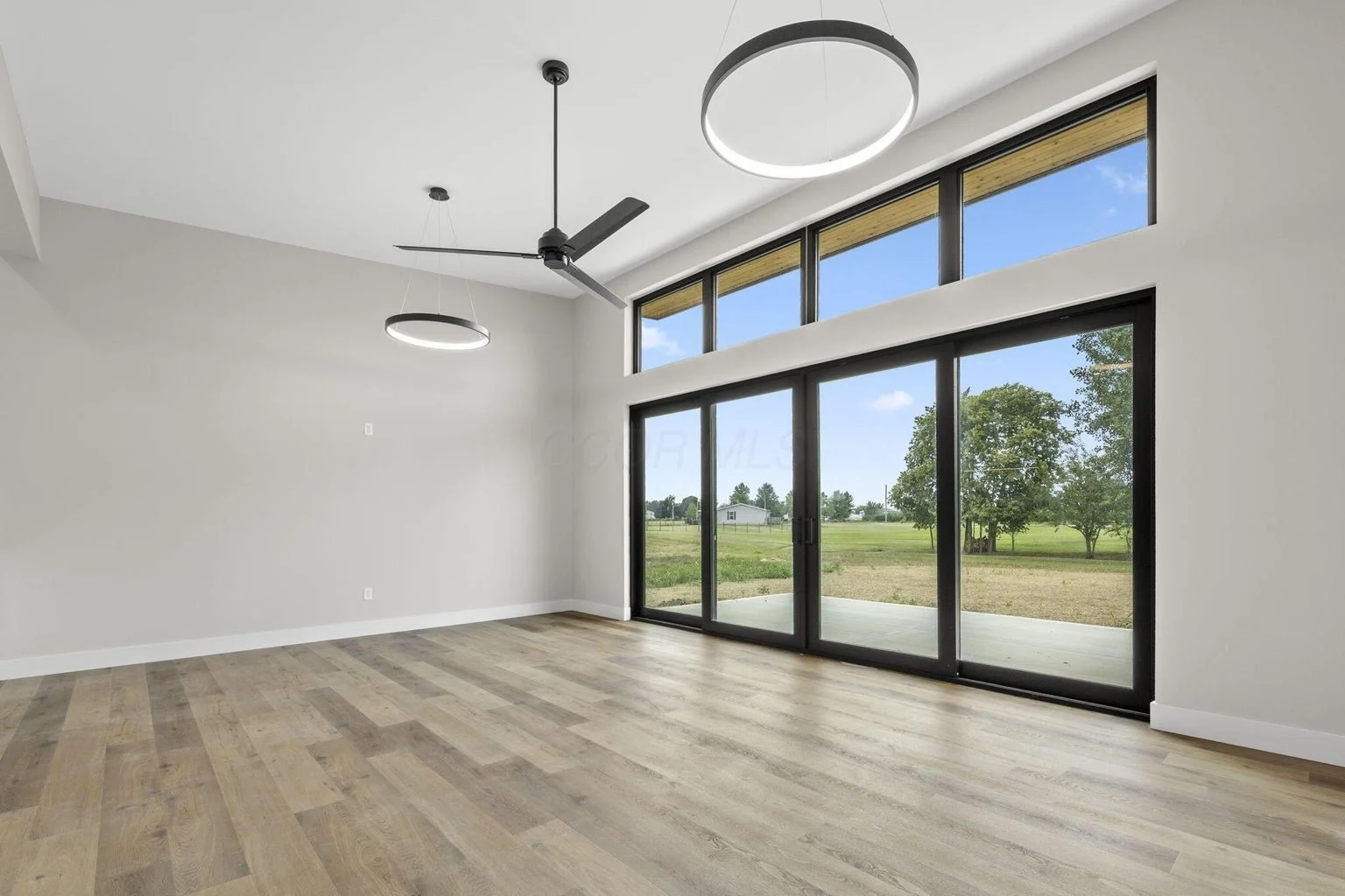 Empty modern living room with large floor-to-ceiling windows showing outdoor green landscape, light wood flooring, white walls, ceiling fan, and circular light fixtures.