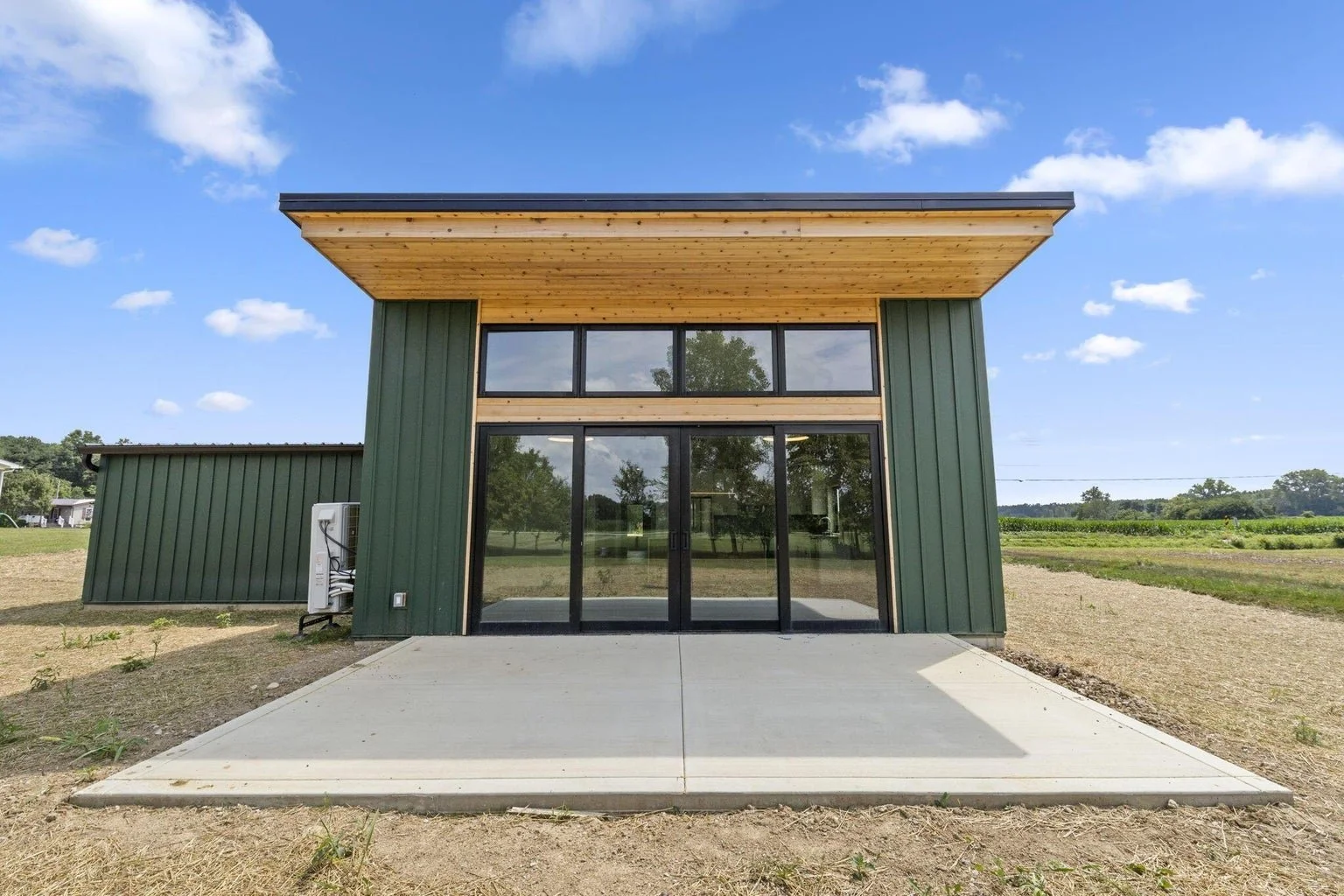 Modern two-story house with green metal siding, large glass sliding doors, and a wooden overhang on the upper level, situated on a concrete patio with open field and blue sky in background.