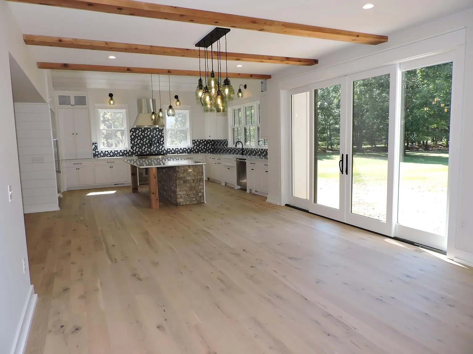 Bright open kitchen with white cabinets, black patterned tile backsplash, wooden beams on ceiling, hanging pendant lights, sliding glass doors opening to a green outdoor yard, light wood flooring.
