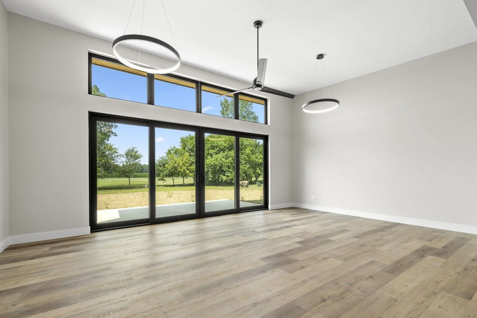 Empty modern living room with large glass sliding doors and windows, light-colored wooden flooring, white walls, ceiling fan, and circular light fixtures, overlooking a green outdoor landscape.