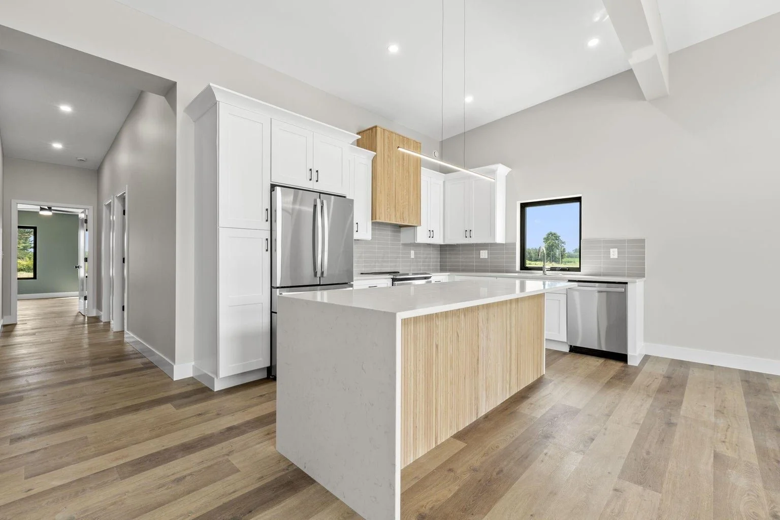Modern kitchen with white cabinets, stainless steel refrigerator, light wood accents, gray tile backsplash, island with white countertop, large window, hardwood floors, and recessed lighting.