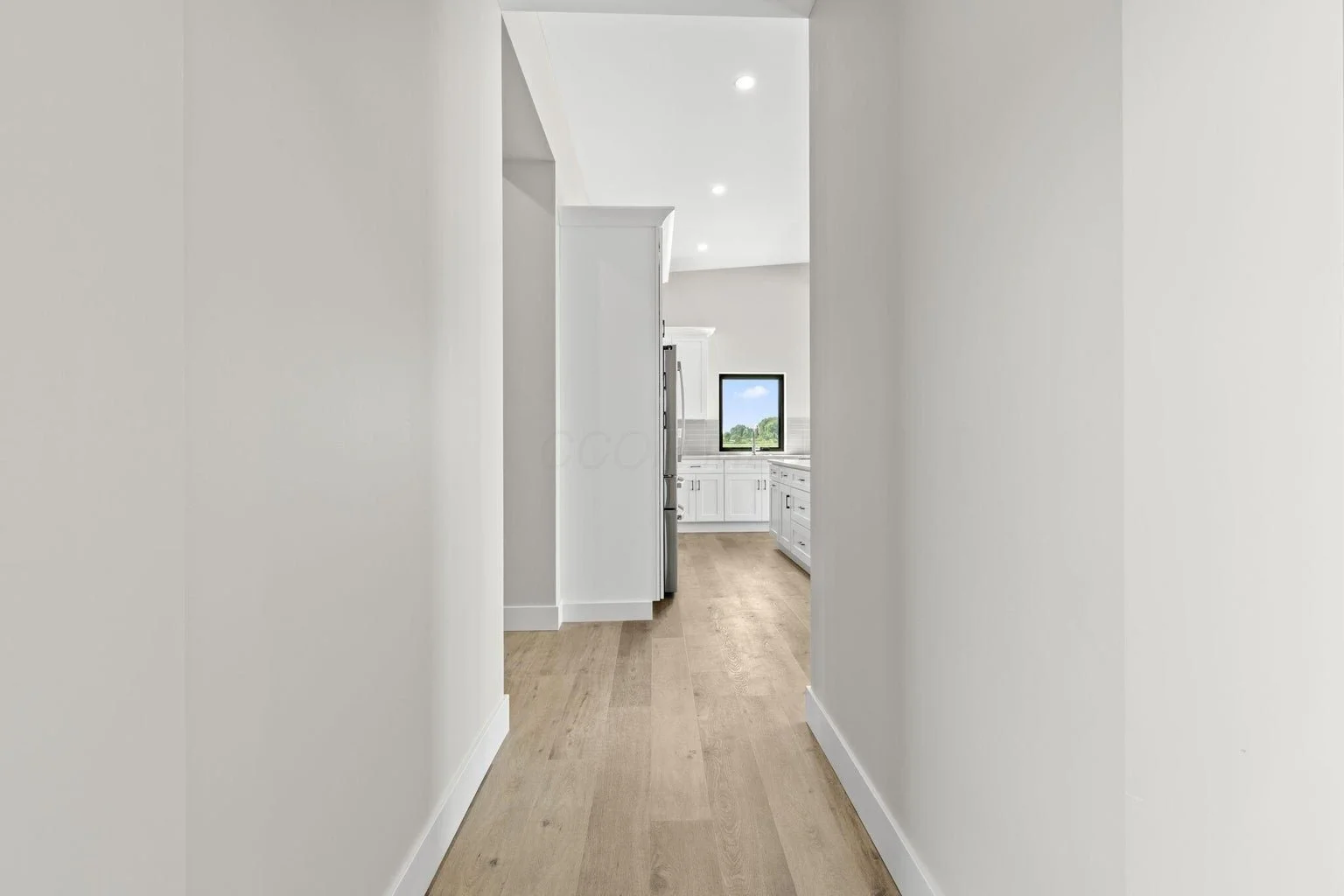 Empty hallway leading to a kitchen with white cabinets and a window showing a green landscape and blue sky.
