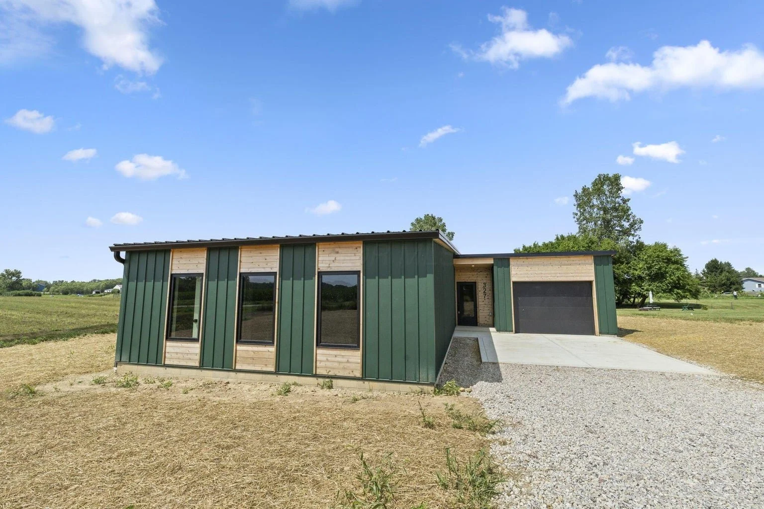 Modern house with green and wood exterior, three tall windows, and a garage on a spacious lot under a blue sky.