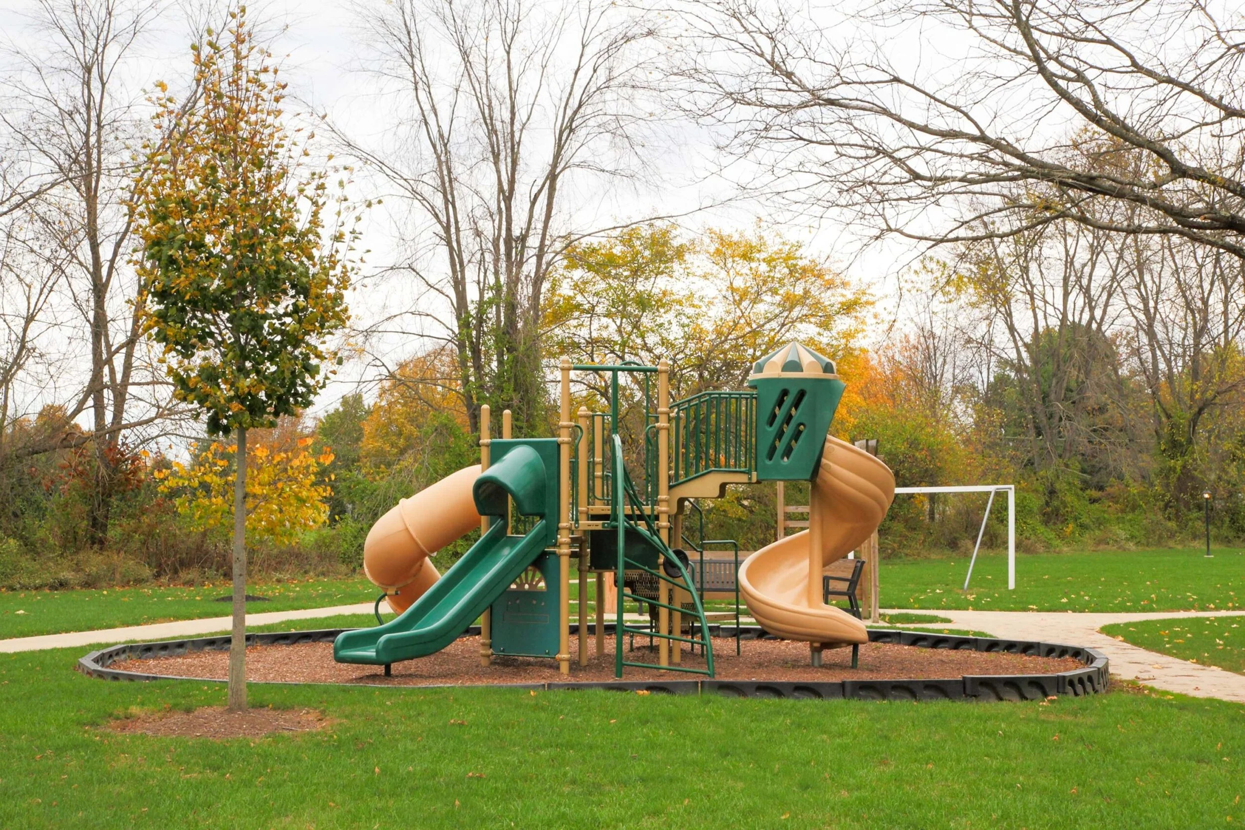 Playground with slides and climbing structures in a park surrounded by trees with autumn foliage.