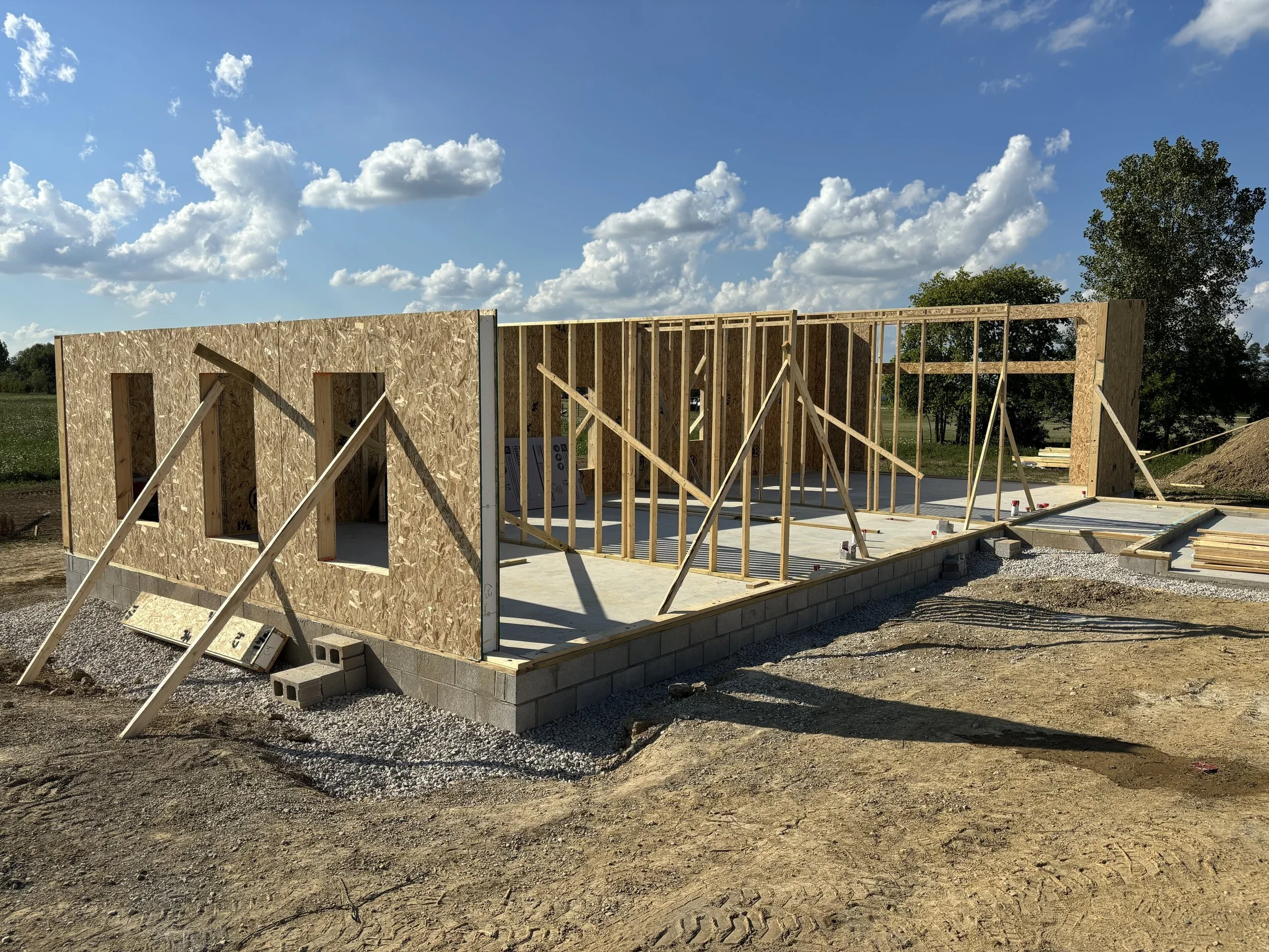 Construction site with wooden framing and OSB walls for a building, set on a concrete foundation with gravel around it, under a partly cloudy sky.
