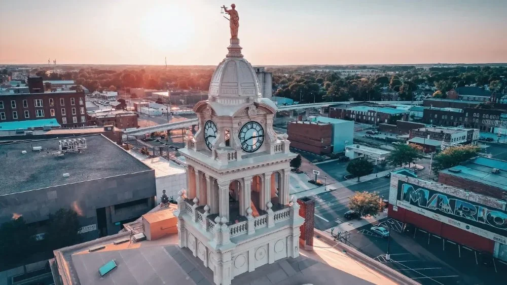 A tall clock tower with a cupola and a statue on top, showing a time of 3:25, in a small town at sunset.