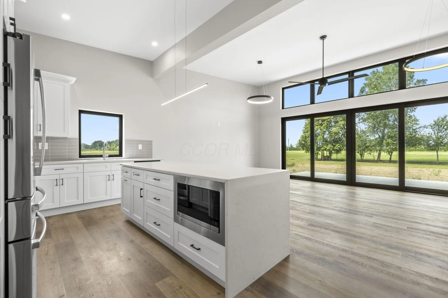 Modern kitchen with white cabinets, a large island, stainless steel appliances, wood flooring, and large windows showing green trees outside.