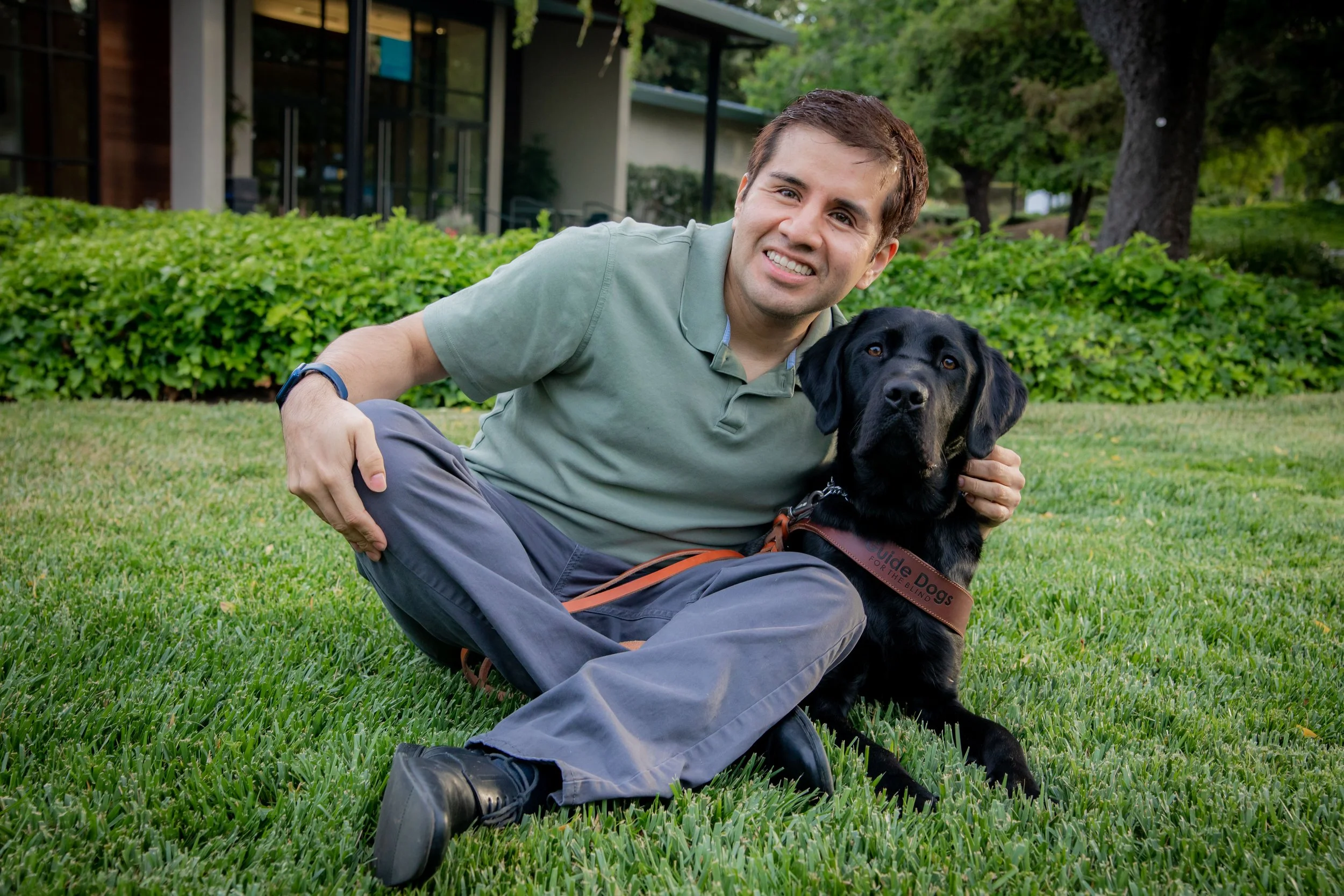 A smiling Latinx man sitting in the grass with his black lab guide dog.