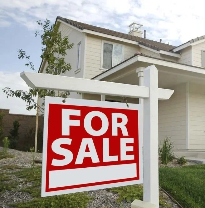 A house with beige siding and a brown roof, with a large white "For Sale" sign in the front yard.