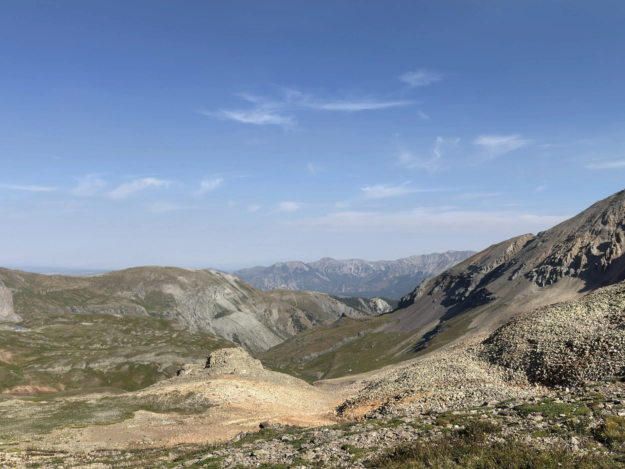 A photo from Telluride, CO: a Scenic mountain landscape with rolling hills, rocky slopes, and distant mountain peaks under a blue sky with a few clouds.