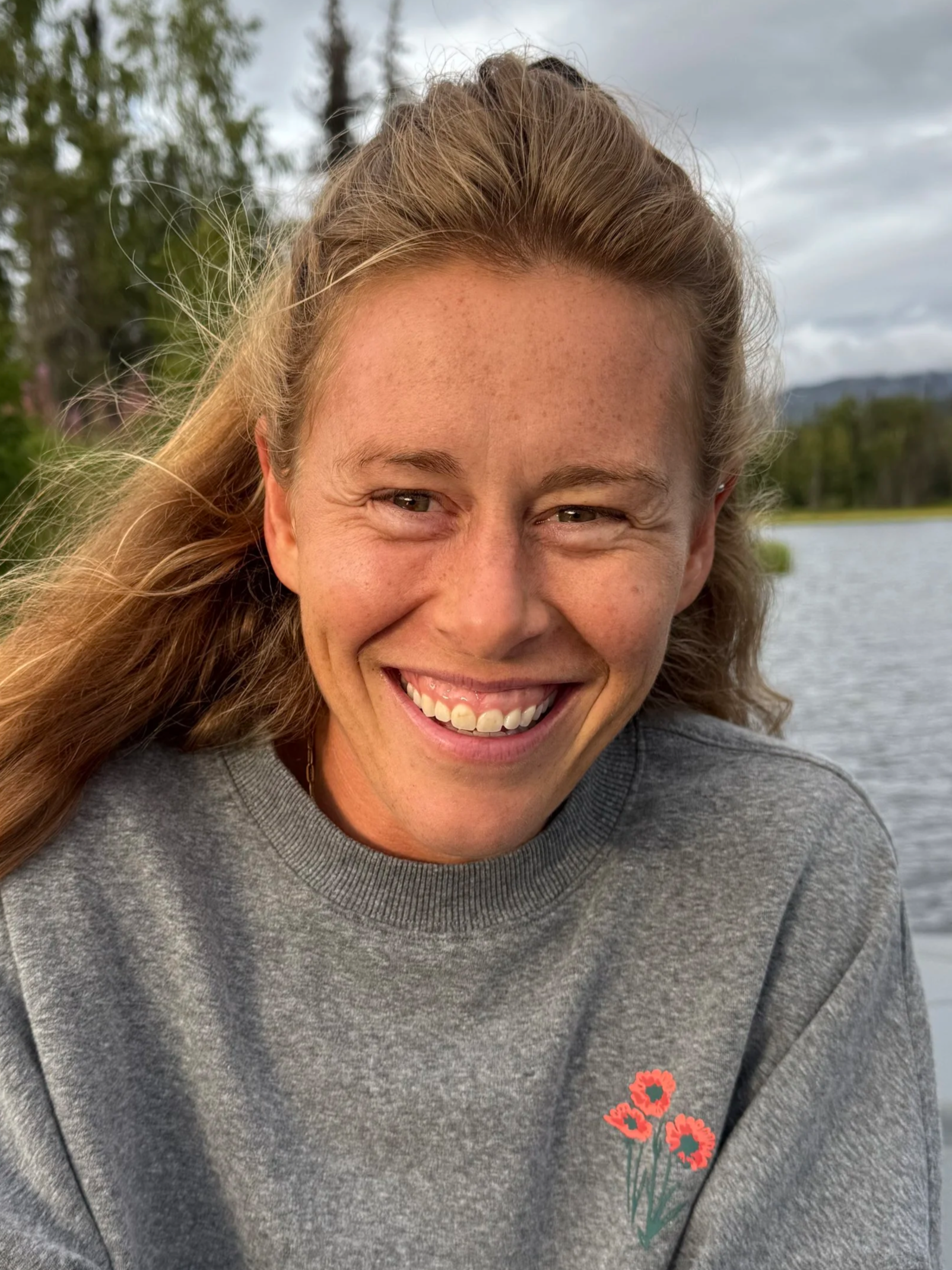 Close-up of professional athlete, mental performance coach, and author Hillary Allen, wearing a gray sweatshirt, outdoors near water with trees and cloudy sky in the background.