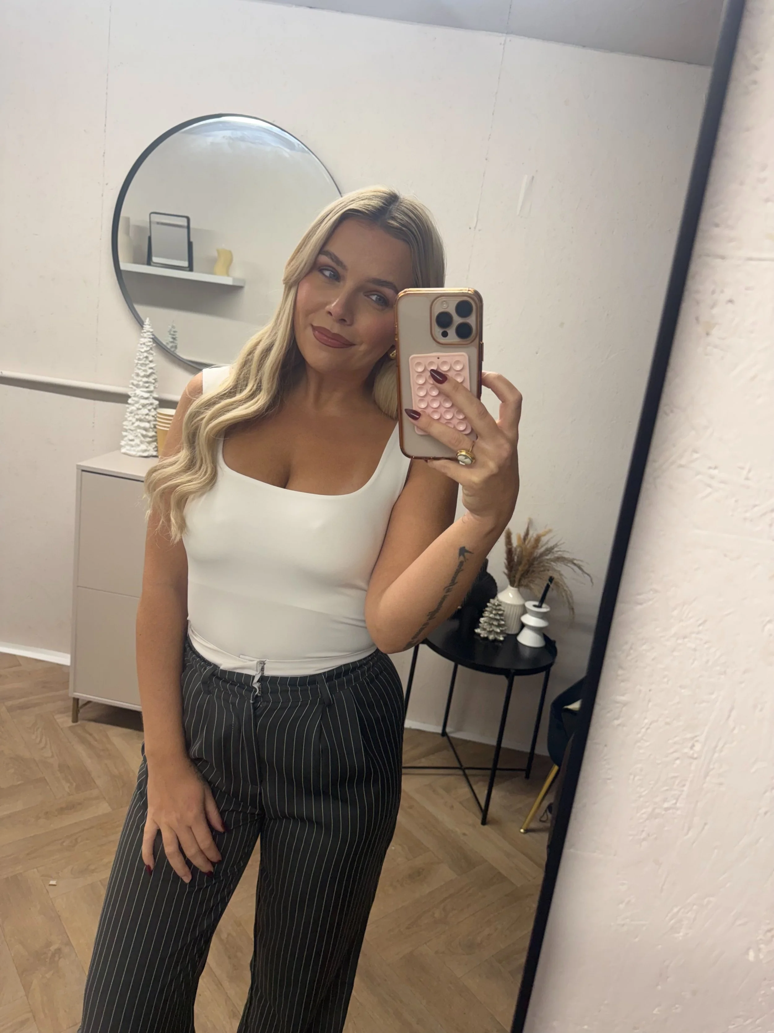A woman taking a mirror selfie in a room with minimalist decor, wearing a white sleeveless top and striped pants.