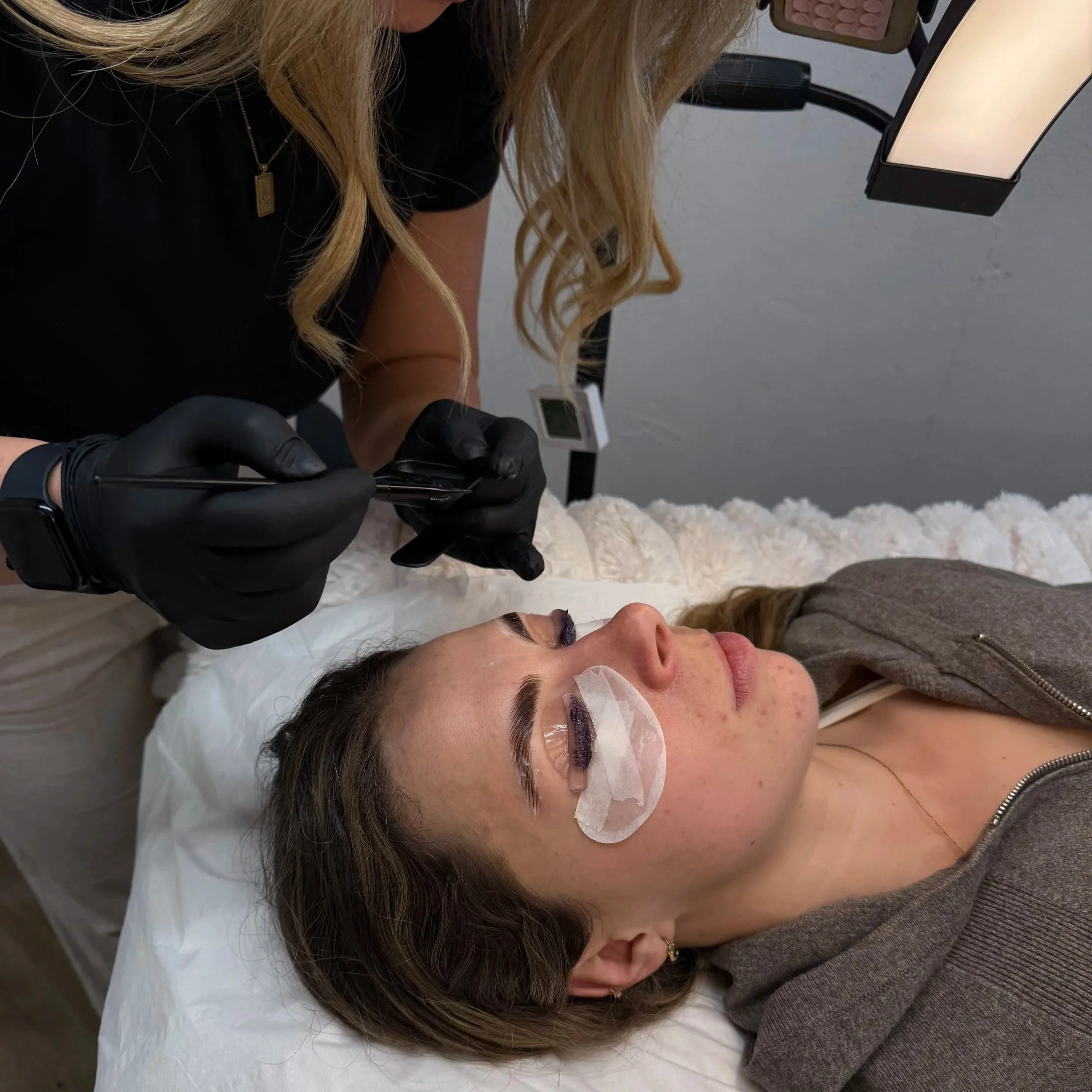 A woman lying on a bed receiving a cosmetic eyelash extension treatment from a technician wearing black gloves.