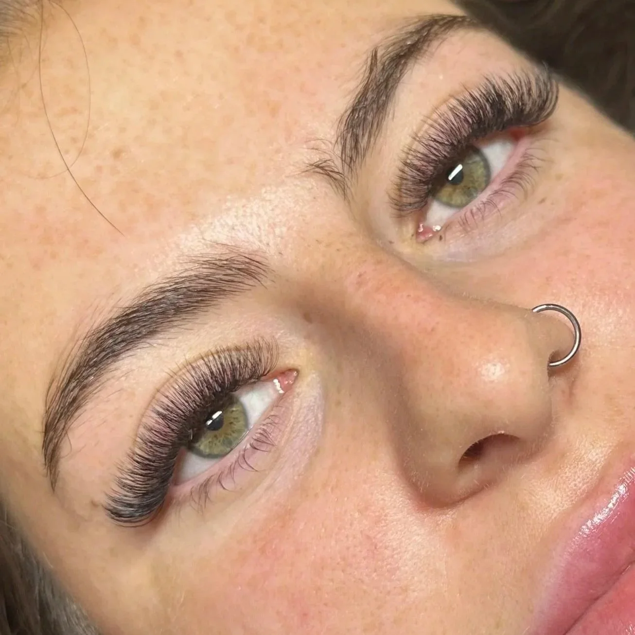 Close-up of a woman's face showing her green eyes, eyelash extensions, well-groomed eyebrows, and a nose ring.