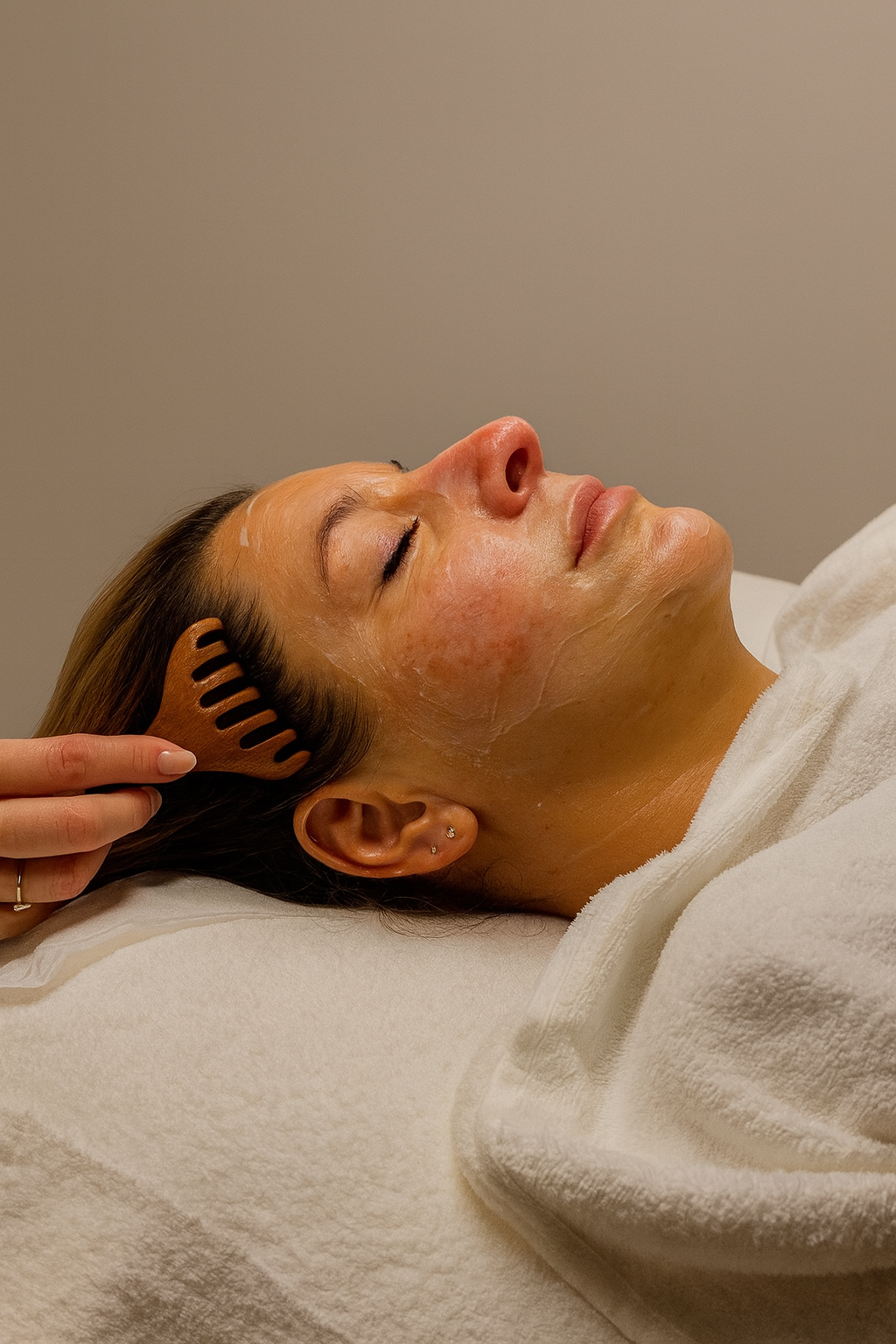 Woman lying with closed eyes receiving a facial massage, with a wooden comb being used in her hair.