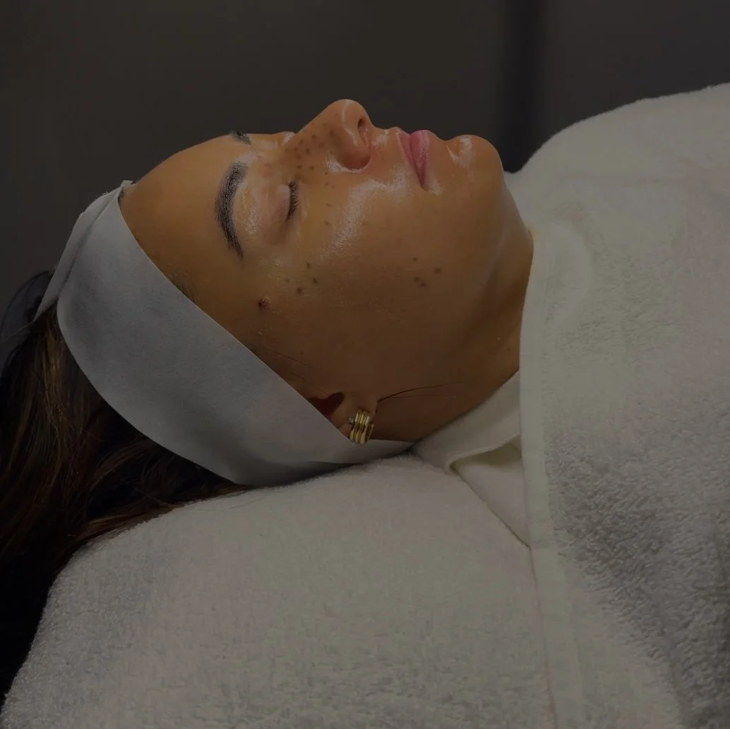 A woman lying down with her eyes closed, undergoing a facial skincare treatment, with a white headband and a white towel covering her chest.
