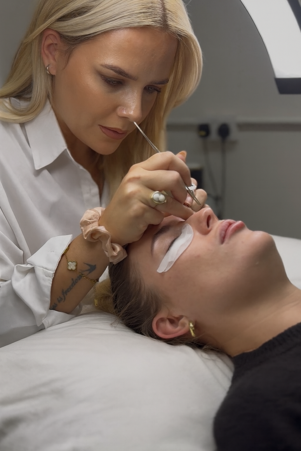 A woman receiving a cosmetic injection under her eye while lying on a bed, with a technician preparing to administer it.