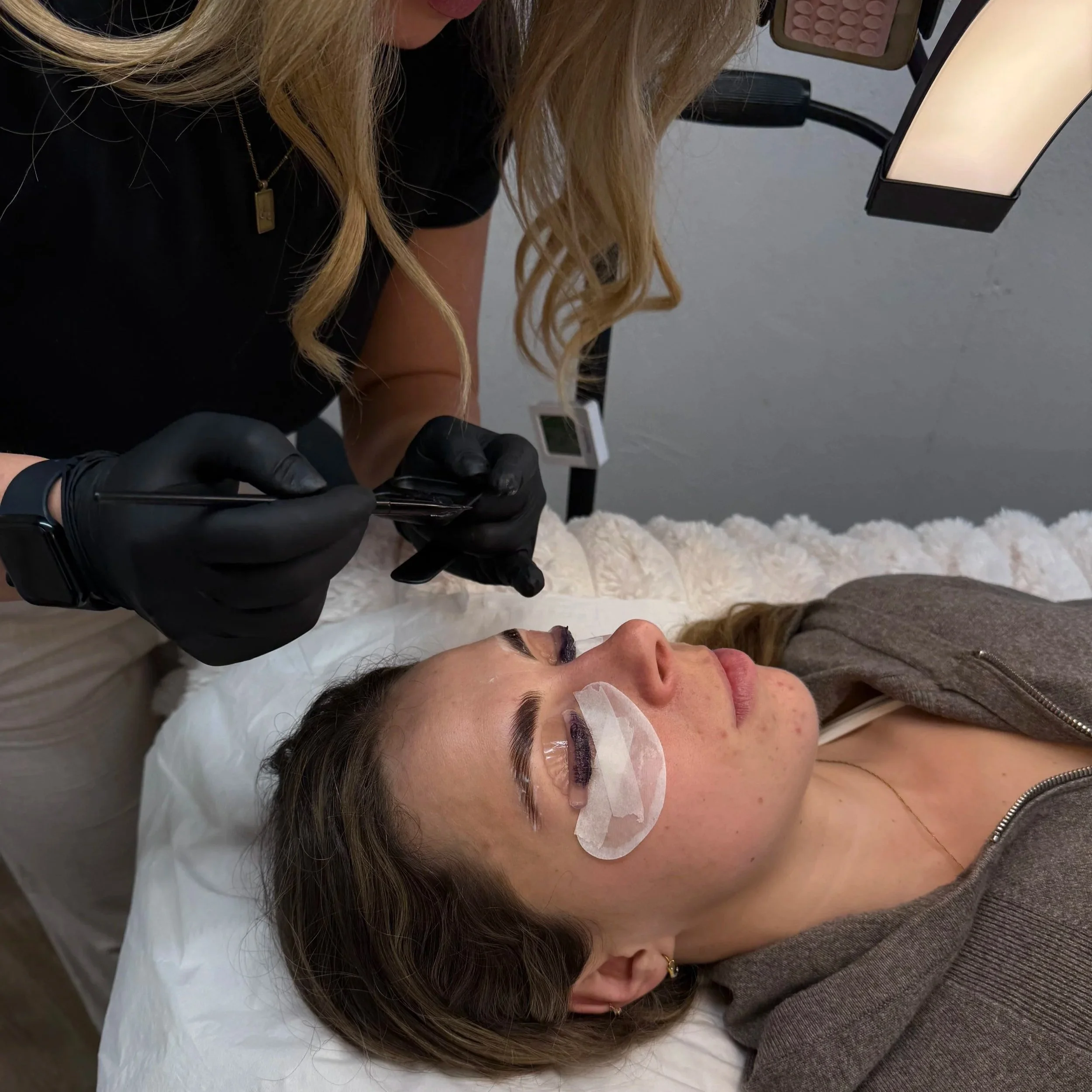 A woman lying down with an eye patch, receiving a cosmetic procedure on her eyebrows from a technician wearing black gloves, in a beauty clinic or spa setting.