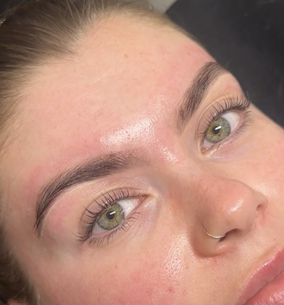 Close-up of a woman's face showing her eyebrows, eyes, and part of her nose with a small nose ring. Her skin appears smooth and slightly dewy.