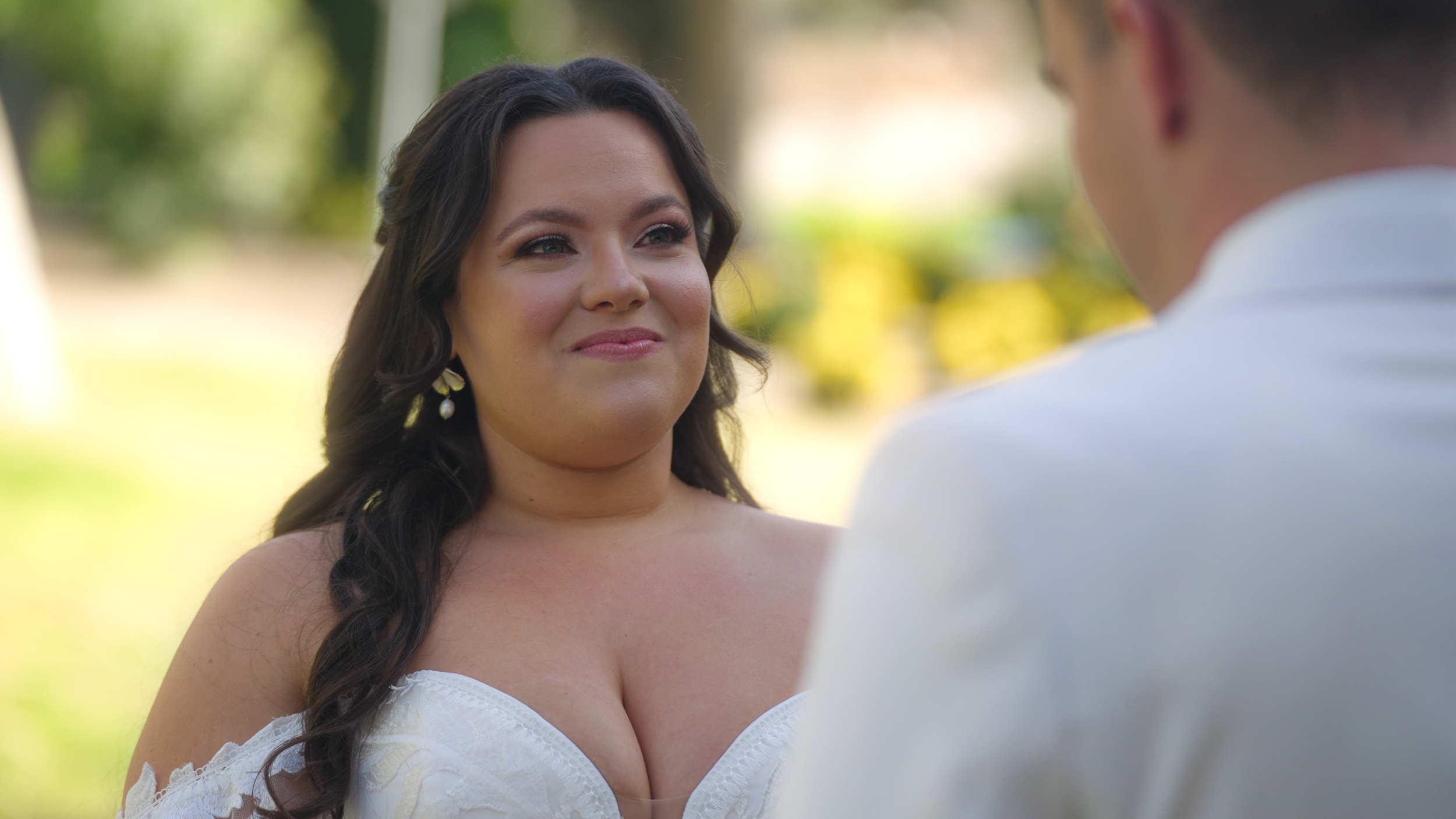 A woman with long dark hair and pearl earrings is standing outdoors, smiling at a man dressed in white, during a wedding ceremony.
