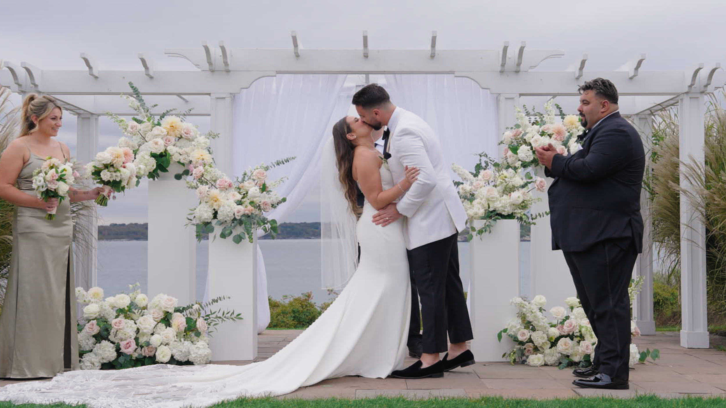 A bride and groom kiss during their outdoor wedding ceremony, with bridesmaids and groomsmen holding bouquets of white and pink flowers, all under a white pergola with a scenic water view in the background.