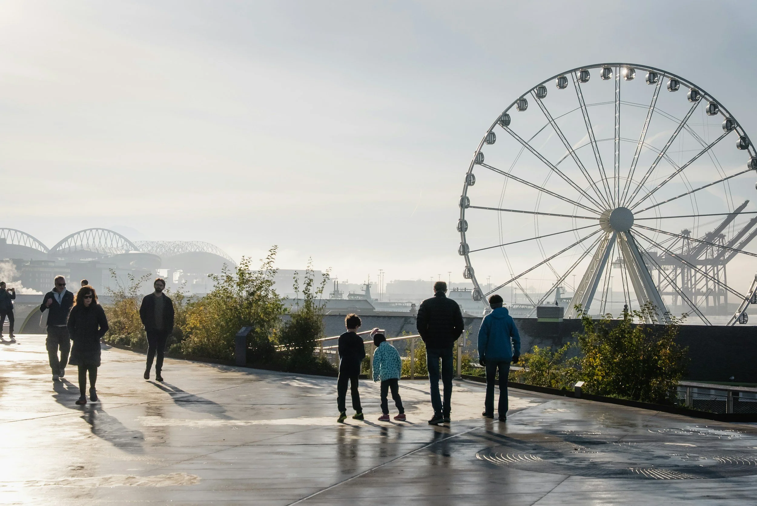 People walking along a waterfront with a large Ferris wheel and other amusement rides in the background.