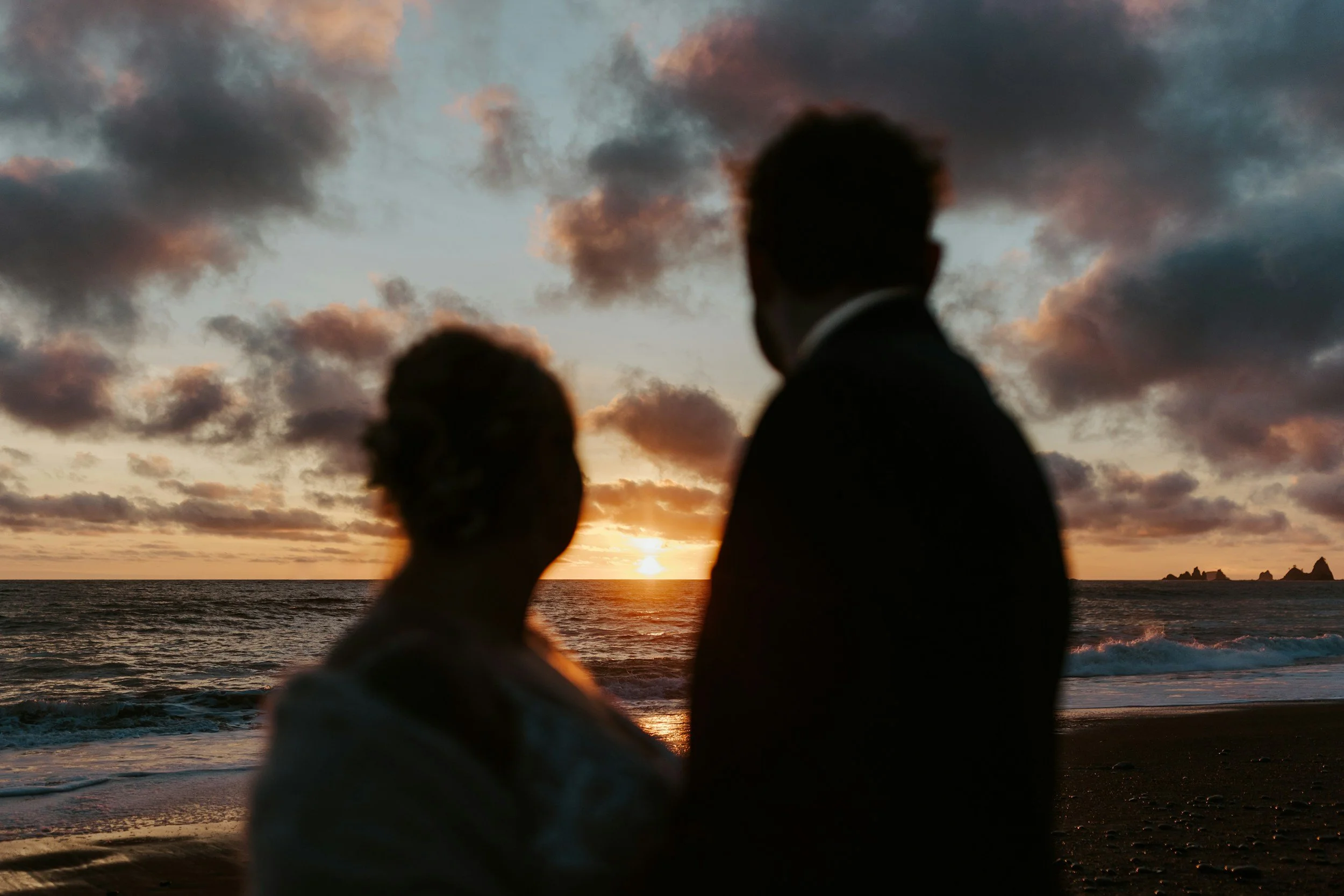 Silhouettes of a man and woman standing on the beach at sunset, facing the ocean with clouds in the sky.