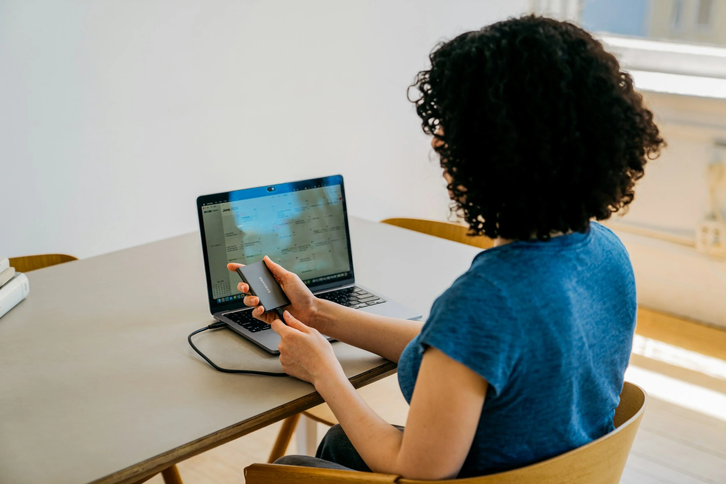 A woman with curly hair wearing a blue shirt sitting at a table with a laptop, holding a portable external hard drive connected to the laptop.