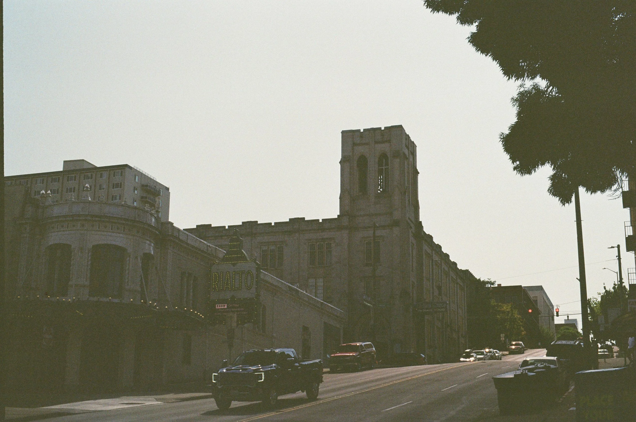 A street view with parked cars, old buildings, and a theater marquee sign that says "RIALTO" visible against a bright sky. Probably in an urban area.