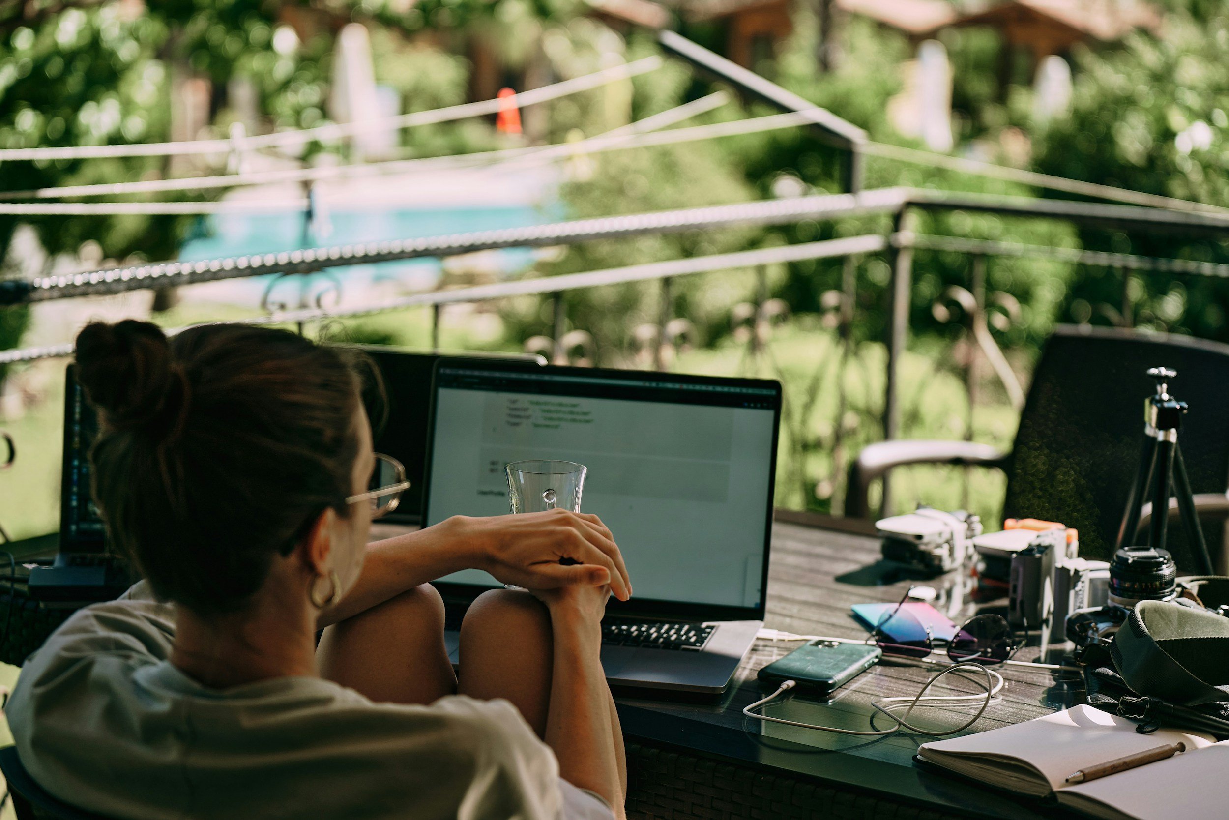 Person working on a laptop at an outdoor workspace with a green, forested background and various electronic devices and equipment on the table.