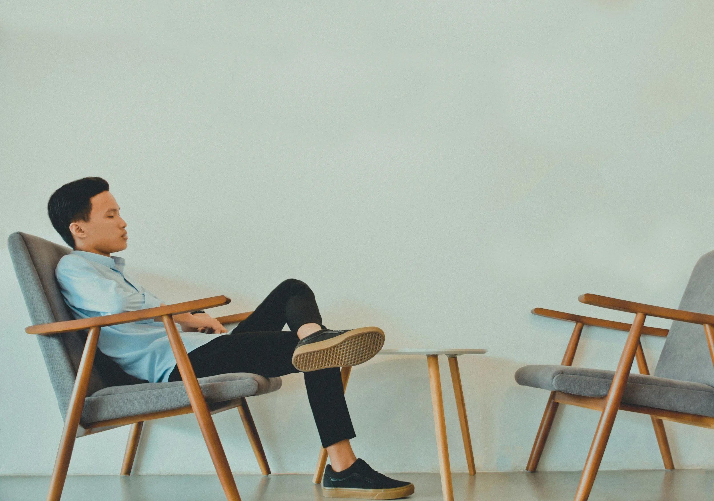A young man relaxing with closed eyes while sitting in a gray cushioned chair in a room with plain light-colored walls, surrounded by empty chairs.