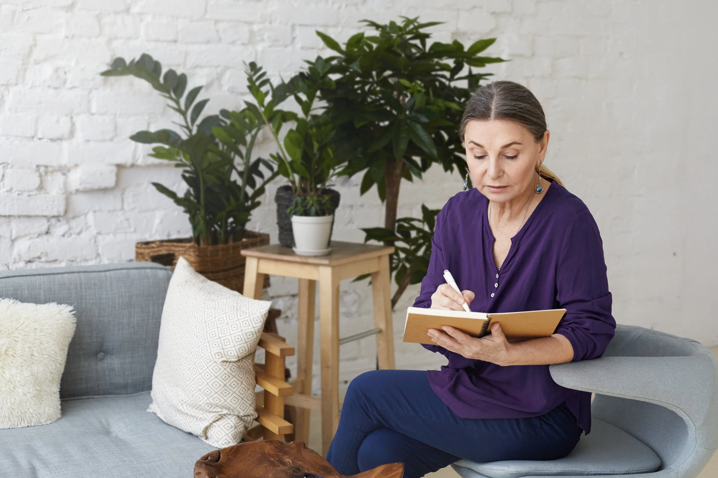 A woman with dark hair pulled back, wearing a purple top, sits on a gray couch writing in a notebook. Behind her are large green plants and a white brick wall, with a small wooden side table with a potted plant.