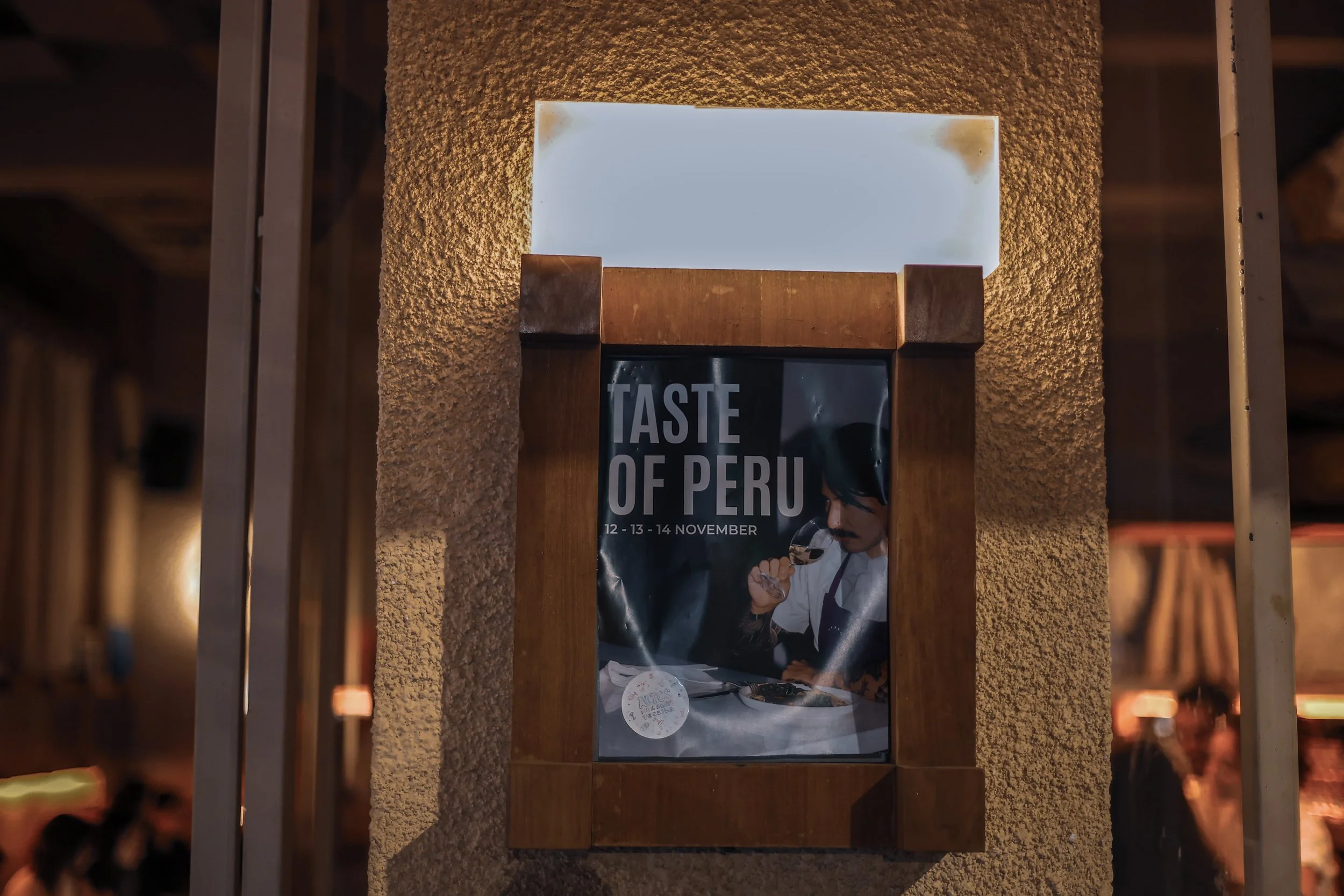 Poster for 'Taste of Peru' event with dates November 12-14, featuring a man tasting food, framed by a wooden border on a textured wall.