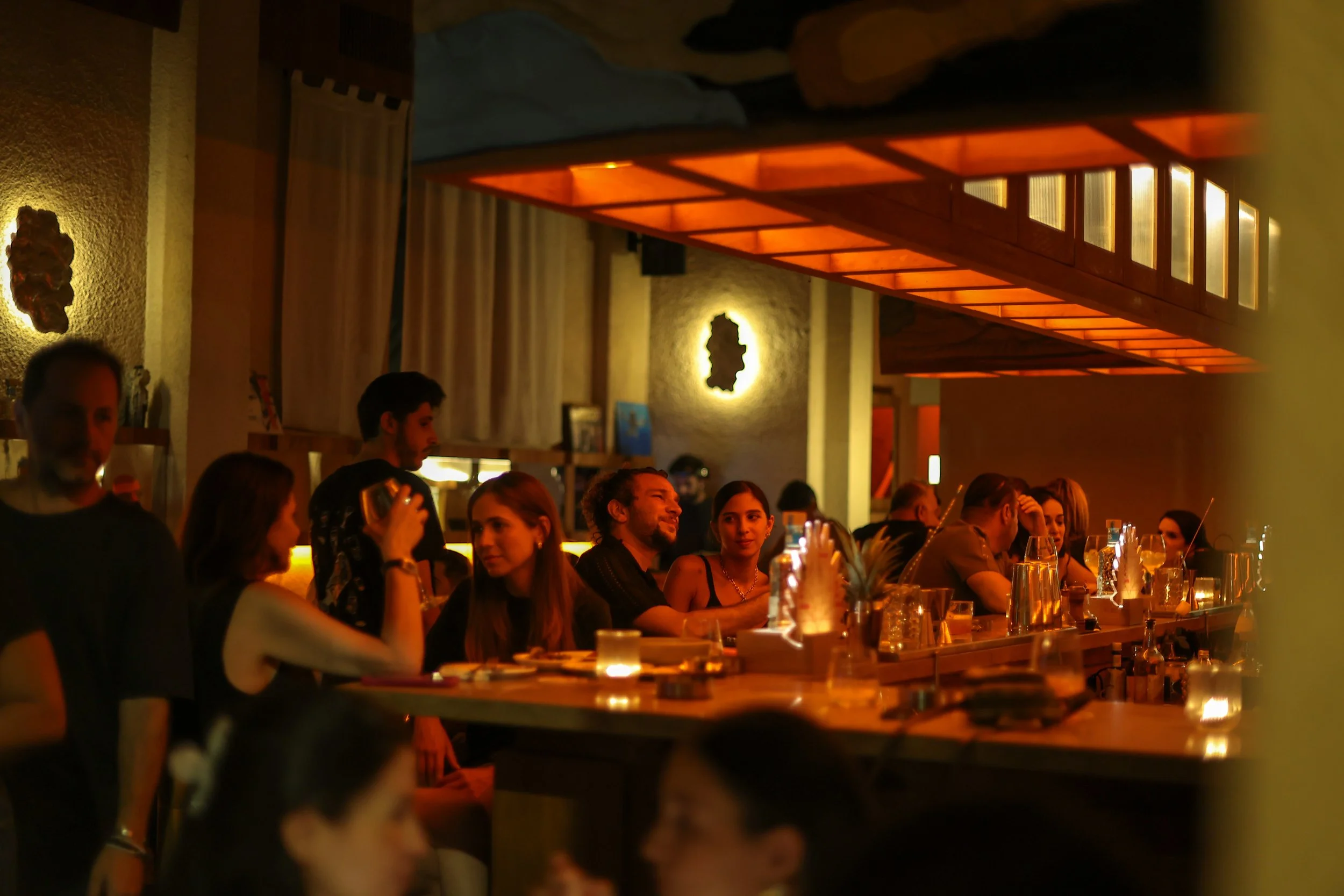 People dining and socializing in a warm, dimly lit restaurant or bar, with a bar counter in the foreground and seating along the wall in the background.