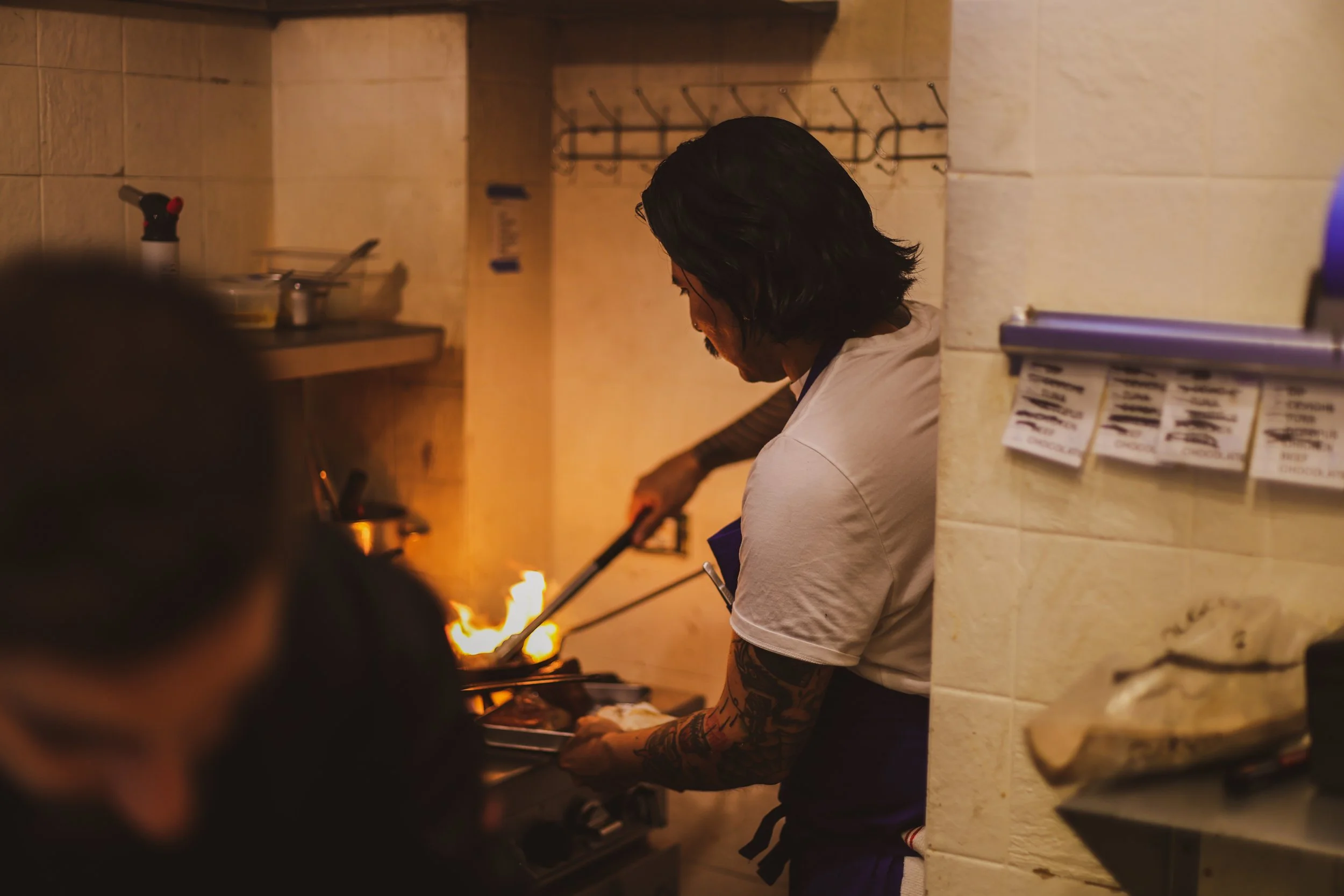 A chef cooking on a stove with a visible flame, wearing a white shirt and apron, in a kitchen with beige tiled walls.