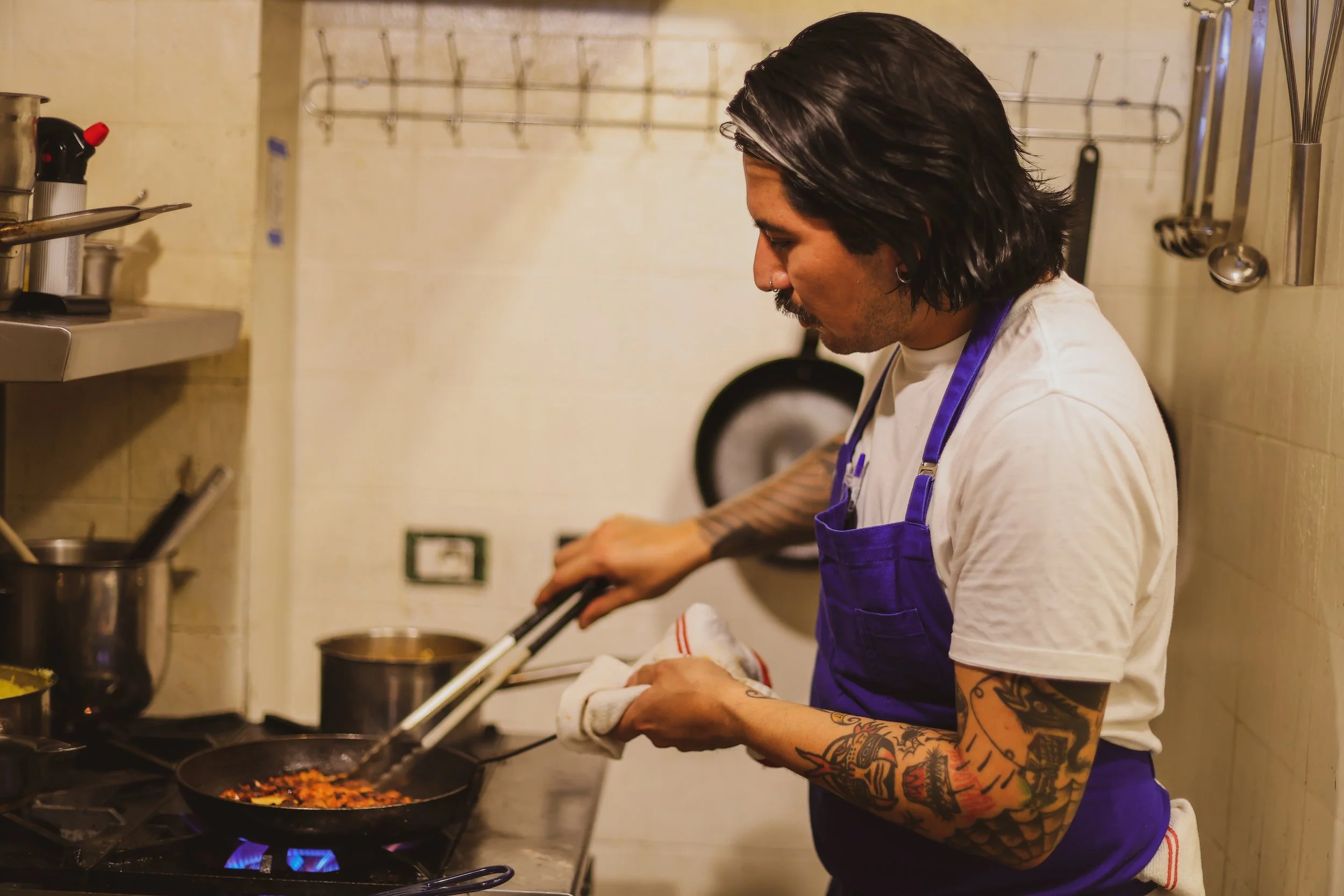 A man with long black hair and tattoos on his right arm, wearing a white t-shirt and a purple apron, is cooking in a kitchen. He is using tongs to stir food in a frying pan on a stove with blue flames. The kitchen has a beige tiled wall, with various utensils hanging and shelves holding pots and tools.