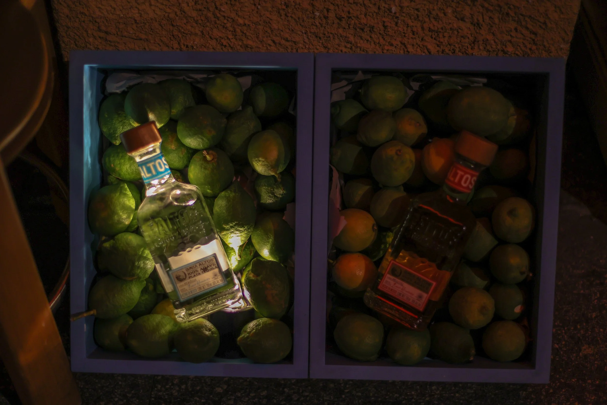 Two boxes filled with green and orange limes, each with a bottle of tequila placed on top.